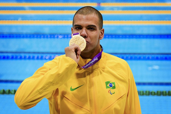 Homem de cabeça raspada, vestindo casaco amarelo, beija medalha de ouro das Paralimpíadas