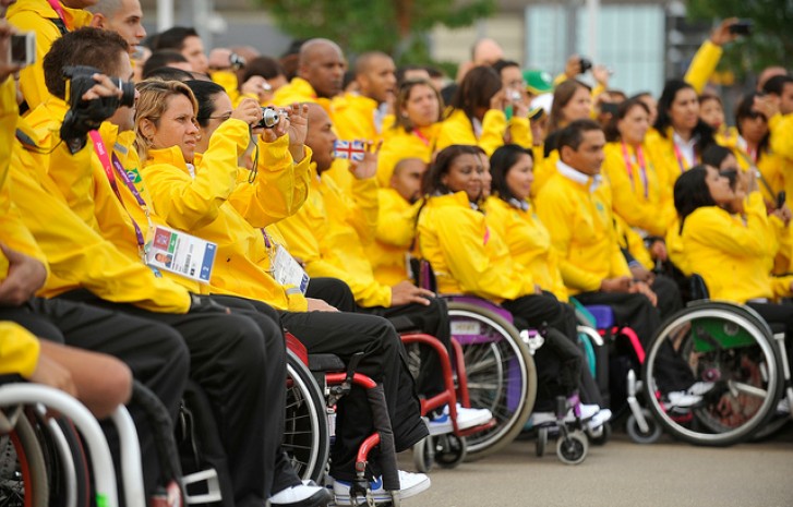 Atletas em cadeira de rodas, vestindo casaco amarelo e calça escura, tiram fotos com máquinas e celulares