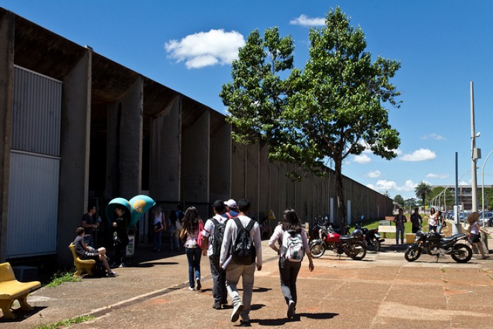 Universitários, com bolsas e mochilas nas costas, andam em direção ao prédio de concreto e horizontal