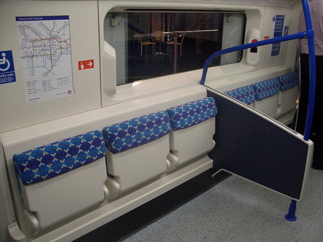 Um vagão de metrô com uma divisória entre seis assentos. Em cima desta divisória, há uma barra de sustentação azul.