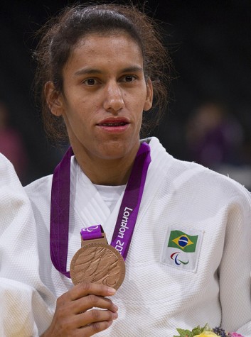 Mulher de quimono branco, com a bandeira do Brasil e símbolo das Paralímpiadas do lado direito, mostra medalha de bronze