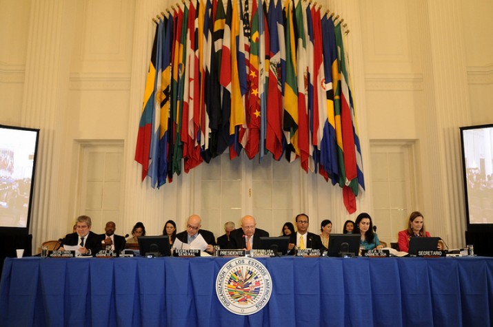 Seis pessoas estão sentadas em mesa coberta por pano azul, com o símbolo da OEA. Na parede, estão penduradas bandeiras de todos os países-membros da instituição