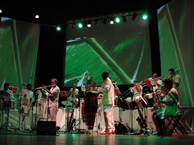 Orquestra de músicos, com instrumentos de sopro e percussão, em palco iluminado por luz verde